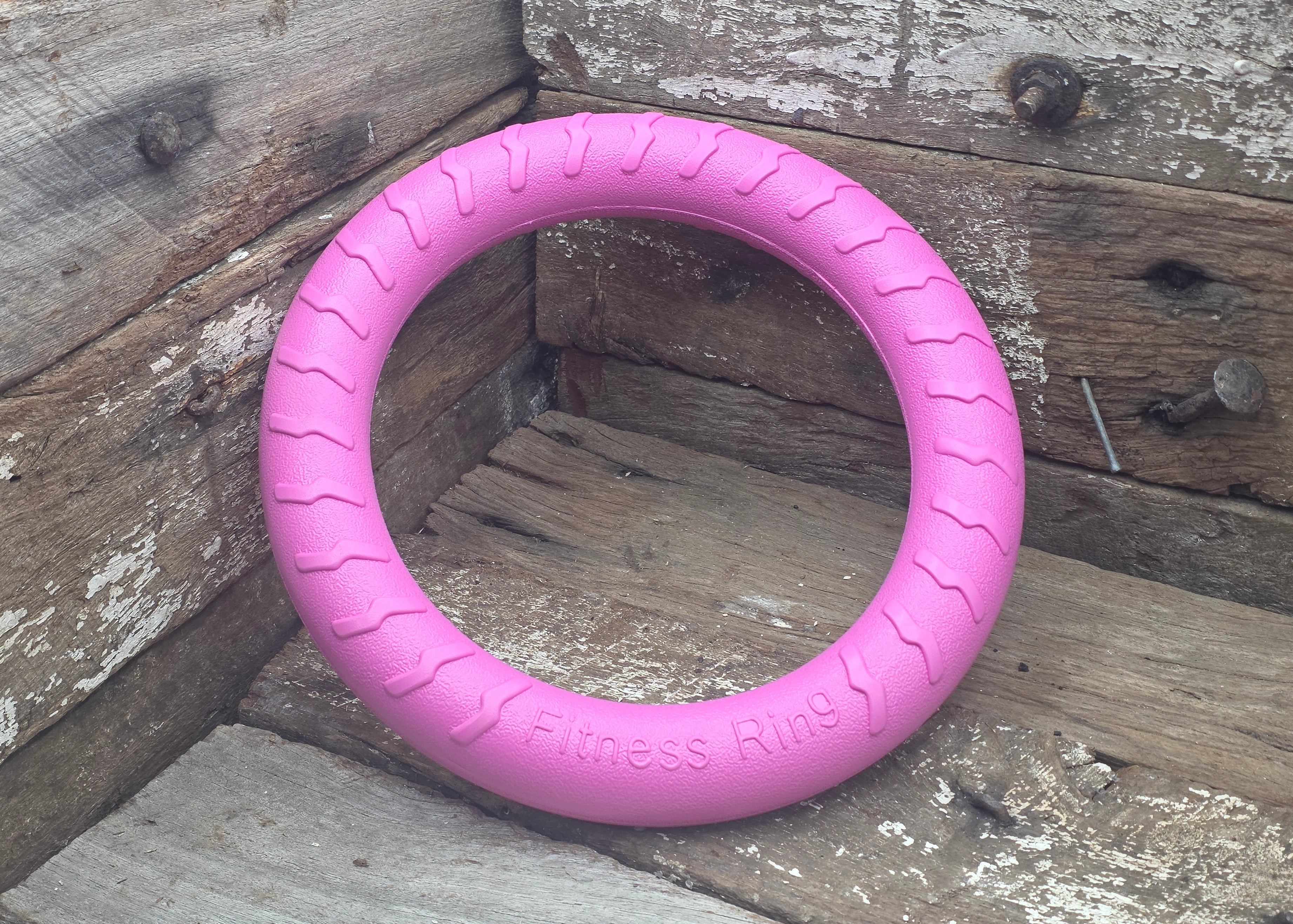 A pink textured circular dog tug toy labeled 'Fitness Rings' resting on rustic wooden boards.