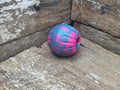 A small textured rubber ball toy with a pink and blue camouflage pattern, resting on a rustic wooden surface.