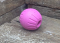 A pink textured rubber ball dog toy with a hollow center, resting on a weathered wooden surface.