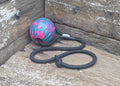 A pink and blue patterned ball attached to a black braided rope toy, resting on a weathered wooden surface.