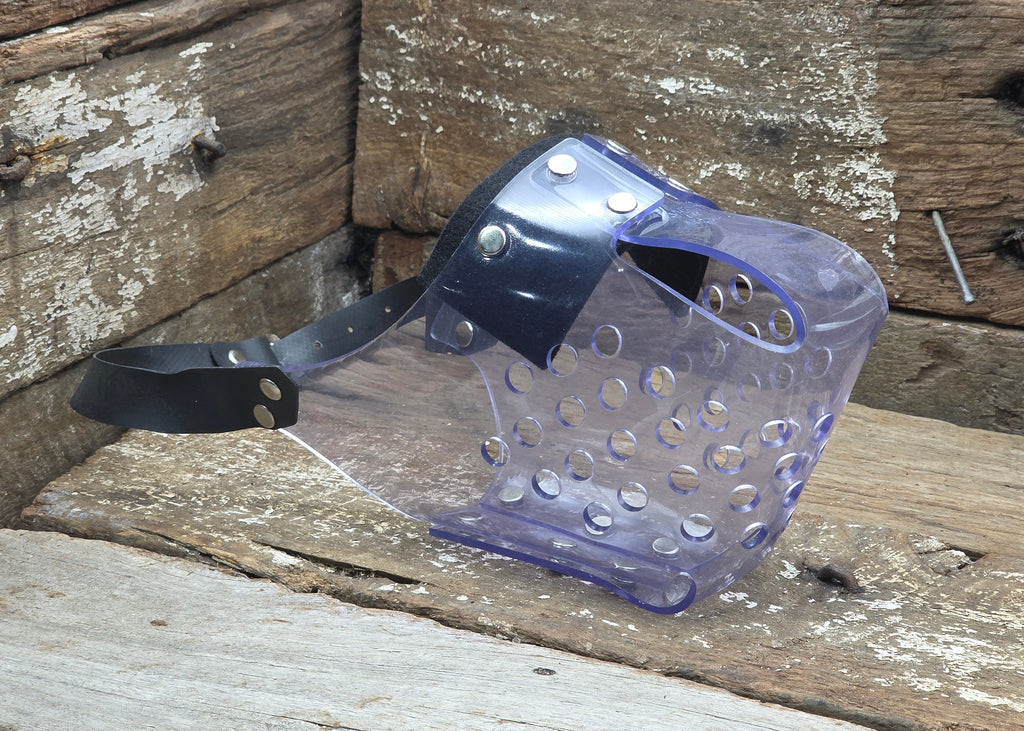 A clear protective face shield with a black head strap and silver metal rivets, resting on a rustic wooden surface.