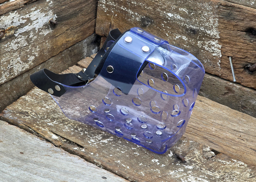 A translucent blue plastic dog muzzle with a black adjustable strap and silver metal rivets, resting on a weathered wooden surface.
