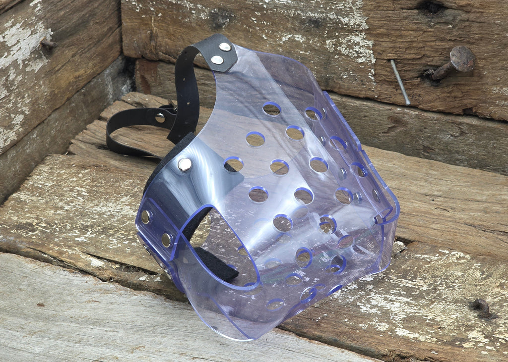 A clear plastic dog muzzle with ventilation holes and a black adjustable strap, resting on a rustic wooden surface.